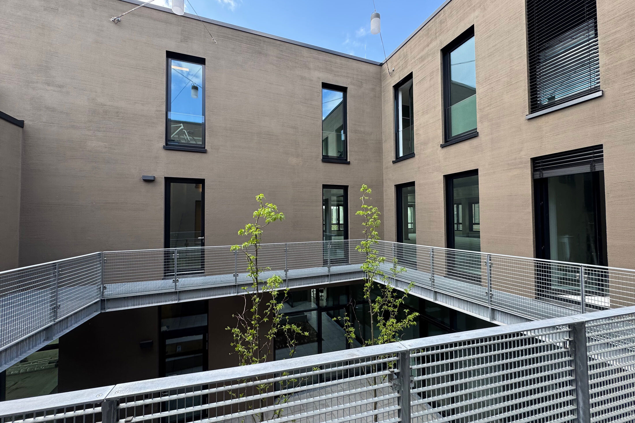 Innenhof moderner Praxis-Neubau mit Metallgeländer, großen Fenstern und jungem Baumbestand.